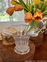 Photo showing clear glass vase with flared petal-like rim and ribbed vertical lines, placed on a brown circular stand with flowers in background.