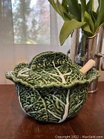 Side view of the green cabbage-shaped ceramic tureen with lid and spoon inside, placed on wooden surface with background showing window and silver vase.