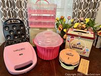 Photo showing two pink Babycakes cupcake makers (one without latch), Nostalgia Electrics Cakepop maker, pink circular cupcake holder with clear lid, and Nostalgia Cakepop storage box with instructions.