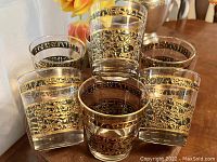 Photo showing seven mid-century drinking glasses with gilt floral and scroll pattern bands around the top. Some fading on the gold gilt decoration is visible.