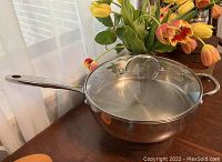 Fry pan with glass lid on dark wooden table next to flowers, showing full pan and handles.