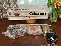 Overall view of the Cuisinart rotisserie box and contents on a table showing the motorized rotisserie unit still sealed in plastic packaging, with the box behind it showing some wear and damage.