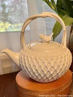 Side view of white ceramic teapot showing textured trellis pattern and bamboo-style handle with bird finial on lid.
