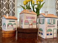 Three vintage ceramic shop house canisters displayed with a background of yellow tulips and neutral curtains. Shows size variation and details of windows, doors, and roofs.
