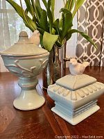 Front and side view of the set showing the taller pedestal container with leaf motifs and the smaller square container, both topped with white bird figurines.