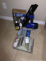 Full view of the lot showing both nailers, boxes of nails, and oil bottles on tiled floor.