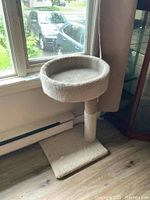 Front angled view of cat stand showing round elevated play area, scratching post and carpeted base near window.