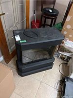 Black electric fireplace with glass front placed on tiled floor near door, showing plug and cord on top.