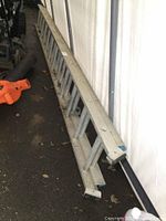 Full view of folded 20 ft aluminum extension ladder on the ground next to a white tent wall, showing minor dirt and wear.