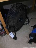 Front angle view of black portable metal fan placed on the floor beneath a wooden shelf.