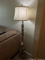 Full view of the floor lamp showing beige fabric shade and wood-look turned base next to a sofa.