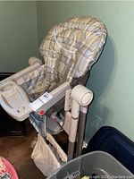 Full view of the Graco high chair with padded plaid cushion, plastic tray, and fabric storage basket below.