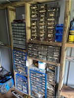 View of seven metal and plastic hardware storage bins of various sizes arranged vertically on wooden shelves containing assorted screws, nails, bolts, anchors, and staples.