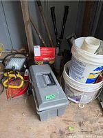 Overview of lot showing grey toolbox, two white plastic pails stacked, garden tools with wooden handles, and Honeywell heater box.