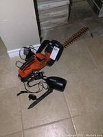 Photo showing the full cordless hedge trimmer with attached charger and light, placed on tiled floor.