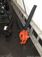 Two Black & Decker electric leaf blowers placed on the ground near a metal ladder, both with black blower tubes and orange bodies.
