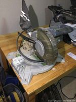 Overall view of the Haus Mann miter saw on a wooden surface showing the saw blade, handle, and dust collection bag.
