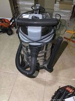 Front view of Maximum 30L stainless steel shop vacuum with hose coiled on top and attachments at the base.