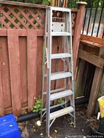Full view of aluminum step ladder standing upright outdoors by a wooden fence.
