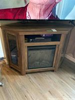 Wide view of wood grain corner TV stand with electric fireplace insert and storage compartments below, holding some media devices.