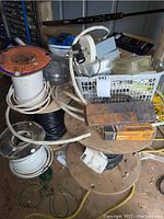 Overview of four partial wire spools on wooden reels, handheld tester, plastic basket with electrical boxes, bucket, and yellow rope on floor
