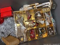 Photo shows a cardboard box containing a plastic tray with many assorted vintage-themed Christmas ornaments such as angels, a truck, animated figures, some with fabrics or faux fur, plus white string lights and stuffed figures with cords.