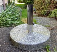 Round marble base umbrella stand placed outdoors on a stone/concrete surface with grass and plants around it.