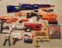 Photo showing multiple Nerf blasters including Modulus and Elite series models plus various attachments like scopes, bipods, and a container of foam darts.