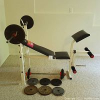 Full view of adjustable weight bench with attachments, deadlift bar positioned alongside, and six varying weight plates placed on floor in front.