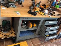 Front view of workbench showing wood top, bench grinder on left, power tools and hand tools on surface, closed drawers and lower shelf.