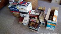 Stacks of paperback and hardcover books in boxes and piles on carpeted floor, assorted fiction and nonfiction titles without visible covers.