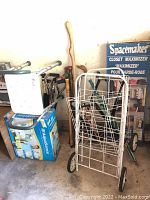 View of grocery cart, wooden canes, boxed raised toilet seat, boxed shower seat, and Spacemaker Closet Maximizer rack box against wall.