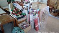 Wide view showing assorted Christmas wreath, gift wrapping papers, gift bows, totes and decorations on and around the chair and table.