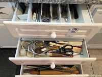 Top drawer with various cleaning brushes, scissors, and kitchen utensils, organized in plastic tray.