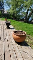 Four flower pots on a wooden deck: two terracotta clay pots and two metal urns, all filled with dirt, set outdoors near grass and trees.