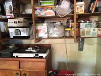 Photo showing white Toastmaster electric griddle on wooden cabinet. Shelving above holds various kitchen supplies and boxes. Black and grey plastic containers next to griddle.