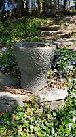 Full view of cylindrical gray cement pot situated on stone steps surrounded by green plants and purple flowers.