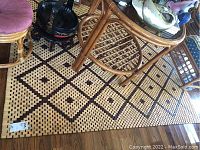 Photo showing the mat laying on a wooden floor with a rattan table on top. Pattern showing wood links in alternating light and dark colors forming diamond shapes with dots inside.