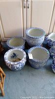 Four ceramic planters in blue and white floral patterns placed on carpet. One planter on a wood base. Various sizes shown.