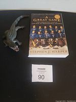 Photo showing the book 'A Great Game' by Stephen J. Harper alongside a plastic toy alligator on a black surface.