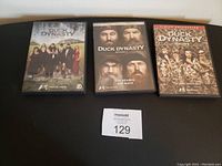 Three DVD cases for Duck Dynasty seasons 1, 2, and 3 placed on a flat surface with a lot number card nearby.