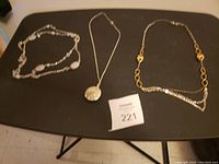Three necklaces displayed on a black surface. Left necklace has multiple silver-tone chains with oval textured beads. Middle necklace has a silver-tone chain with round textured pendant. Right necklace is a gold-tone chain with circular bead accents and linked design.