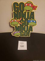 Wall hanging sign with Teenage Mutant Ninja Turtles faces and text 'GO NINJA GO NINJA GO!', yellow background, colorful bandanas on turtles.