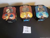 Photo of three Pokemon card holder tin boxes featuring Charizard, Raichu, and Gyarados on honeycomb backgrounds in orange-red, yellow, and blue colors.