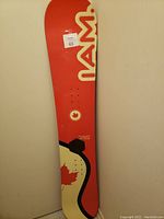 Full view of the red NAM snowboard standing vertically showing logo and maple leaf graphic