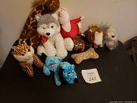 Image showing a variety of stuffed animals arranged on a black surface including a giraffe, husky, pig, lion, cat, blue dog, small bear, and dog bone-shaped plush.