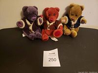 Three stuffed bears in purple, red, and tan, each with a knitted vest, seated against a neutral background with a MaxSold tag number 250 in front.