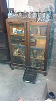 Full front view of the antique glass front wooden cabinet showing three shelves filled with various glass bottles and containers, some mugs, and other small items. Cabinet has a dark wood finish with scratches.