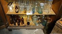 Shelf with various vintage clear and amber glass bottles and jars, some with lids, arranged in two rows on a wooden shelf unit.