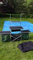 Full view of the assembled portable camping kitchen unit with black countertops, metal frame, green fabric storage compartments, and carrying bag on the grass.